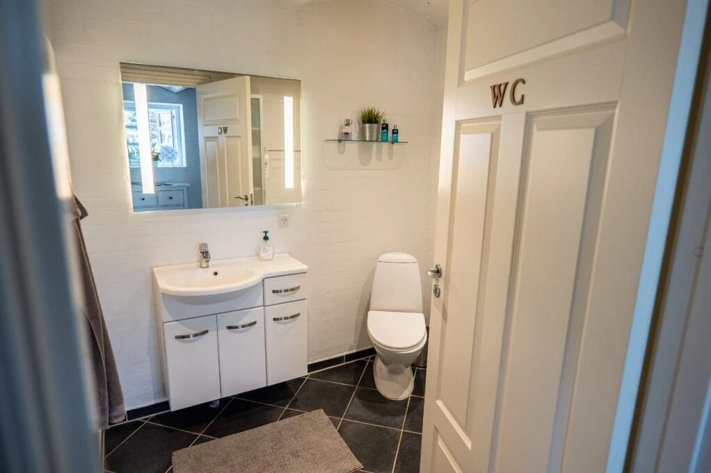 Badeværelse med håndvask, spejlskab og toilet bag en dør med håndmalet WC.