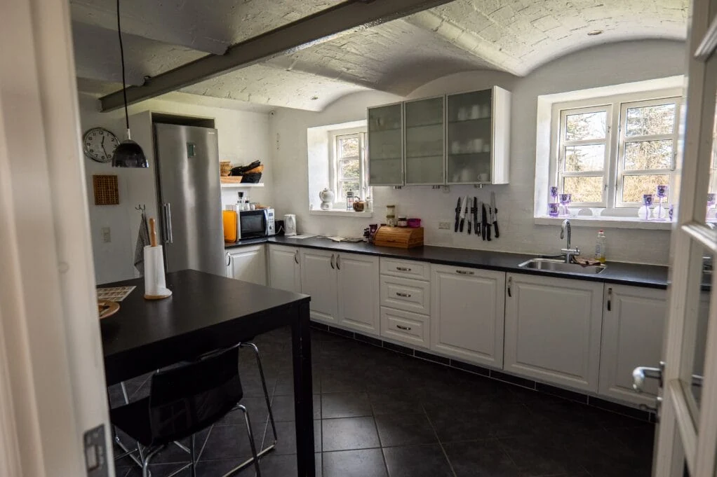 Fælleskøkkenet med hvide låger, mørk bordplade, vask under vinduerne og spisebord med barstole.