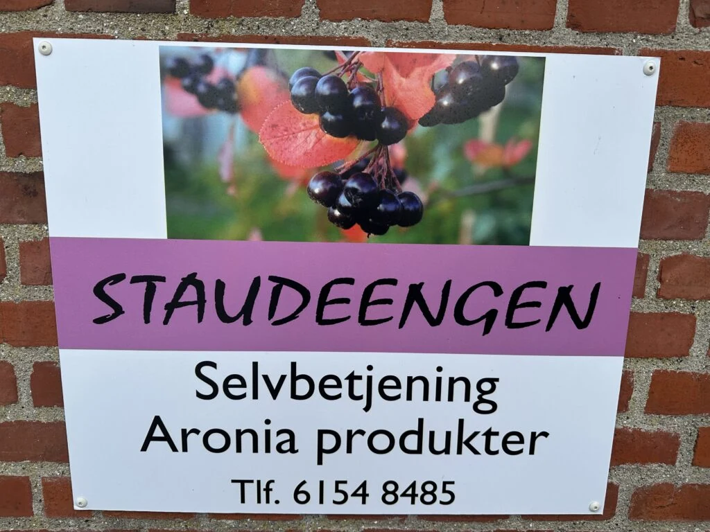 Skiltet på murstensvæggen: Staudeengen — selvbetjening, aronia-produkter, tlf. 6154 8485.